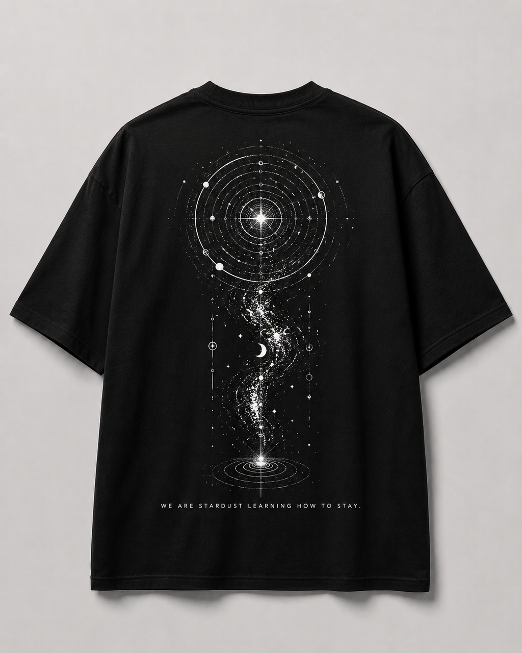 A black short-sleeve t-shirt shown from the back, featuring a large white cosmic graphic of a galaxy, stars, and a wormhole, with the text 'WE ARE STARDUST LEARNING HOW TO STAY.' printed below.