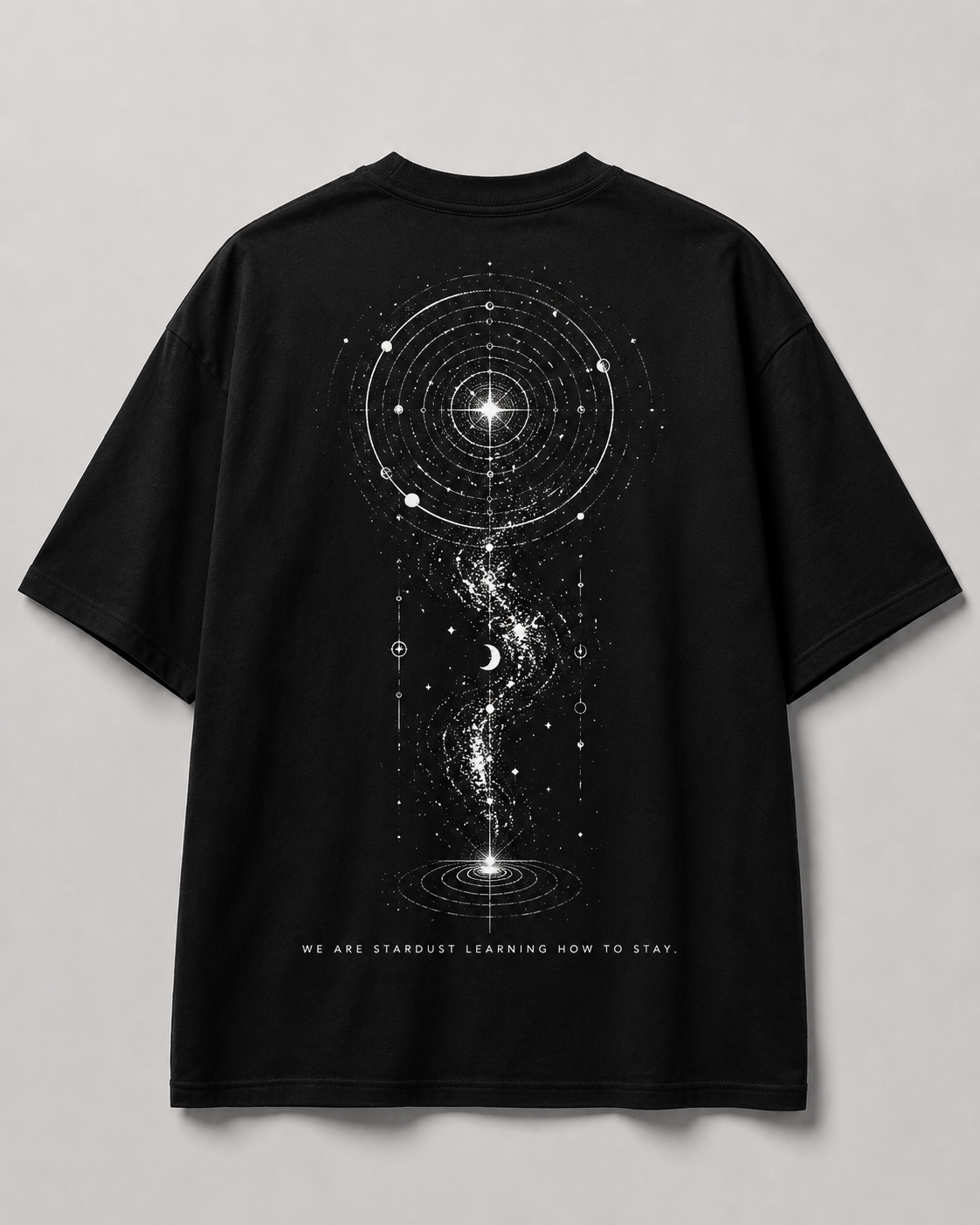 A black short-sleeve t-shirt shown from the back, featuring a large white cosmic graphic of a galaxy, stars, and a wormhole, with the text 'WE ARE STARDUST LEARNING HOW TO STAY.' printed below.